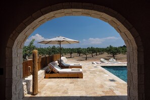 Un espace piscine extérieur agrémenté de chaises longues et d’un parasol, le tout encadré par une arche en pierre donnant sur un vignoble.