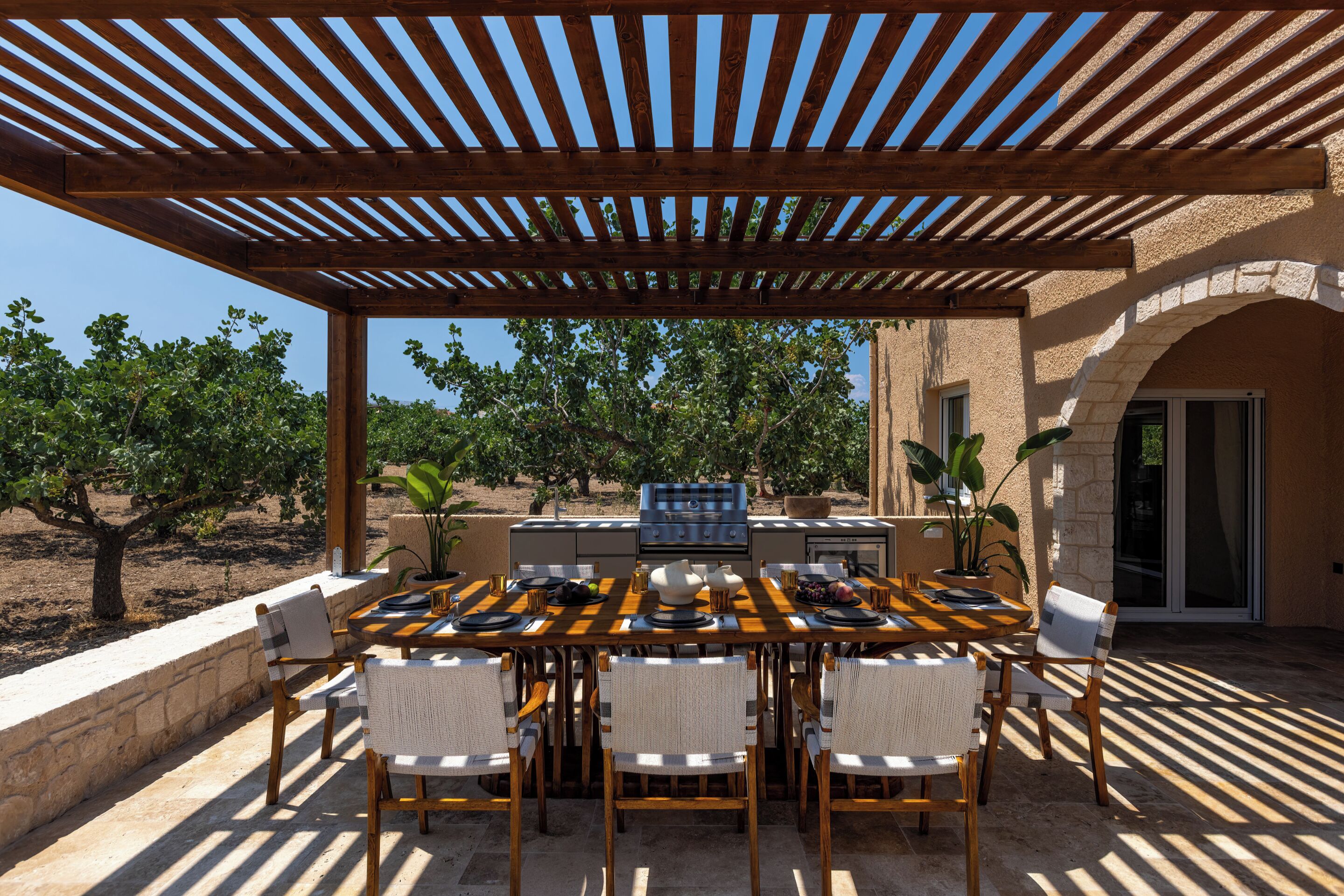 Een terras buiten met een houten pergola, een eettafel gedekt voor vier personen, en een barbecuehoek.