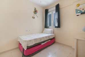 A small bedroom with a single bed, a window with curtains, a wall decoration, and a small table with a lamp.
