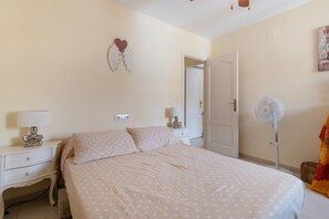 A bedroom with a bed, bedside tables, a wall-mounted fan, and a door leading to another room.