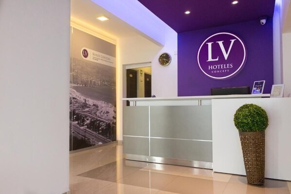 A hotel reception area with a reception desk, a clock, and a poster on the wall. - LV Hoteles (Viña del Mar)