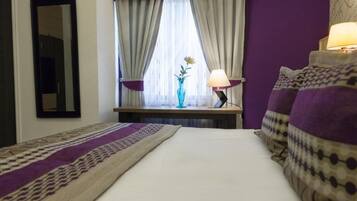 Una habitación de hotel con cama, mesita de noche con lámpara, ventana con cortinas y un florero con flores.