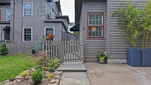 A residential property with a stone pathway leading to a wooden gate, a small garden with various plants, and a grey building with a red-framed window.