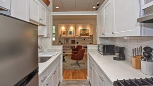 A modern kitchen with white cabinets, a stainless steel refrigerator, a microwave, and a coffee maker. There is a seating area with a brown chair and a rug.