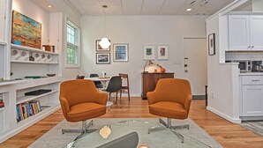 A modern living room with a glass coffee table, two orange armchairs, a dining area with a white table, and a kitchen with white cabinets.