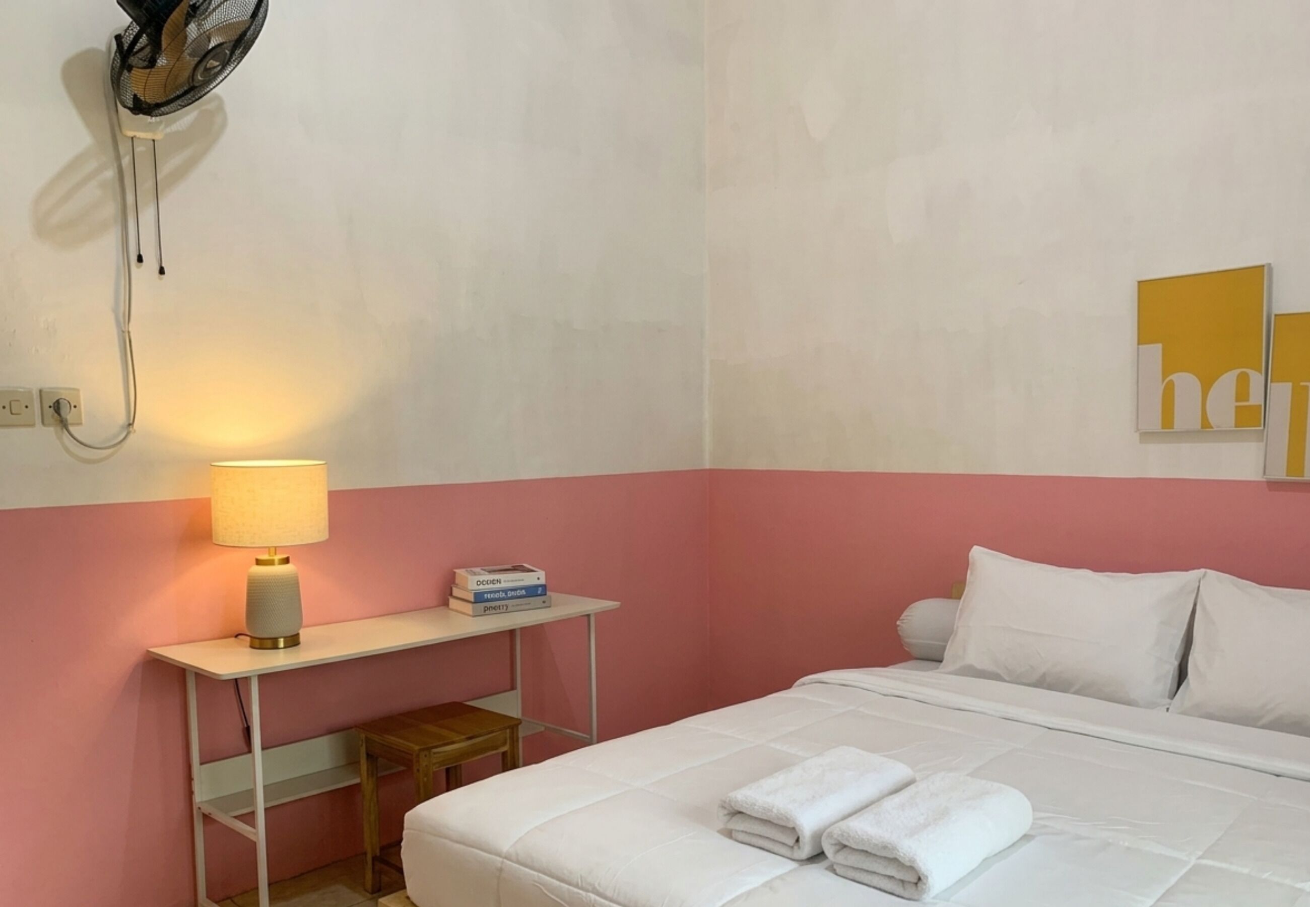 Una habitación de hotel con una cama, una mesita de noche con lámpara, una pequeña mesa con libros y un ventilador de pared.