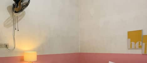 Una habitación de hotel con una cama, una mesita de noche con lámpara, una pequeña mesa con libros y un ventilador de pared.