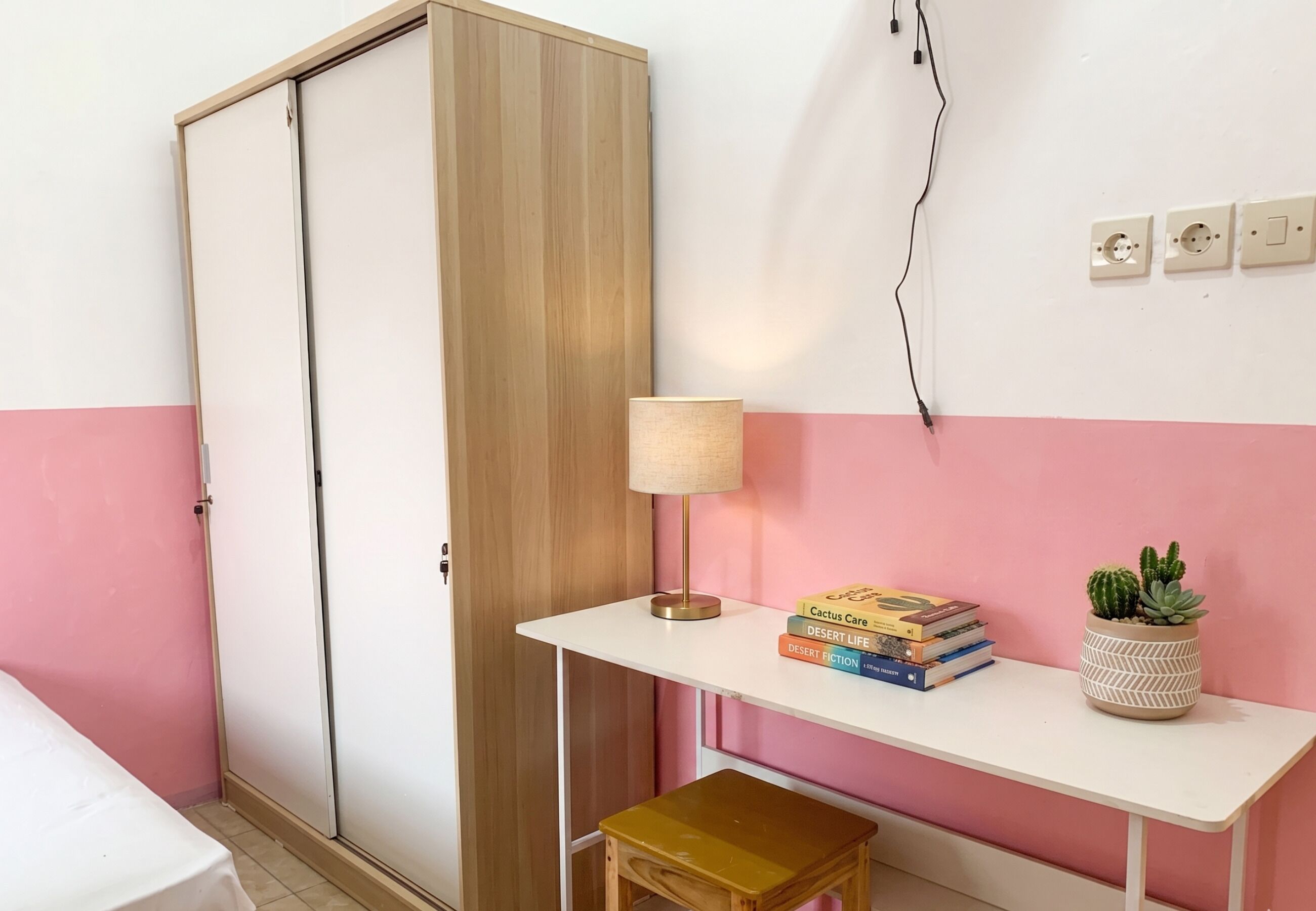 A room with a wardrobe, a desk with a lamp, a small stool, a plant, and books.