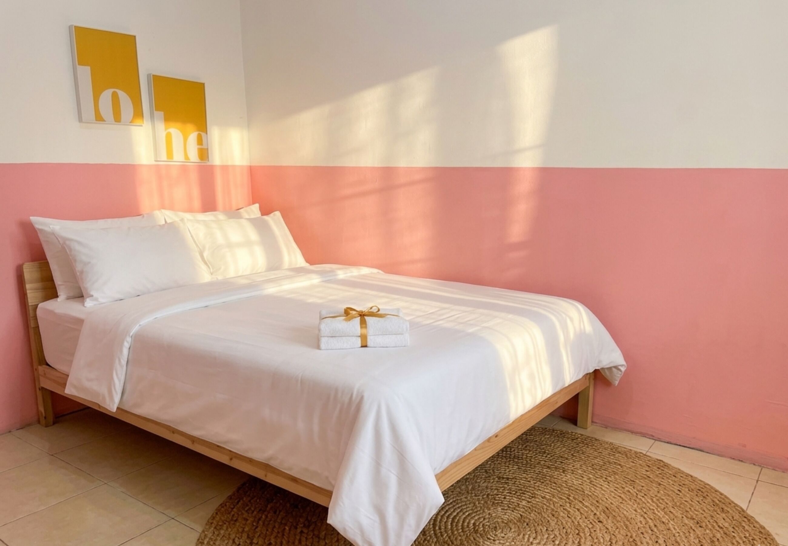 Una cama individual con colchón y almohadas blancos, estructura de madera y una alfombra tejida sobre piso de baldosas.