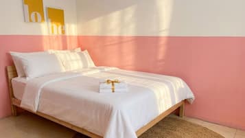 Una cama individual con colchón y almohadas blancos, estructura de madera y una alfombra tejida sobre piso de baldosas.