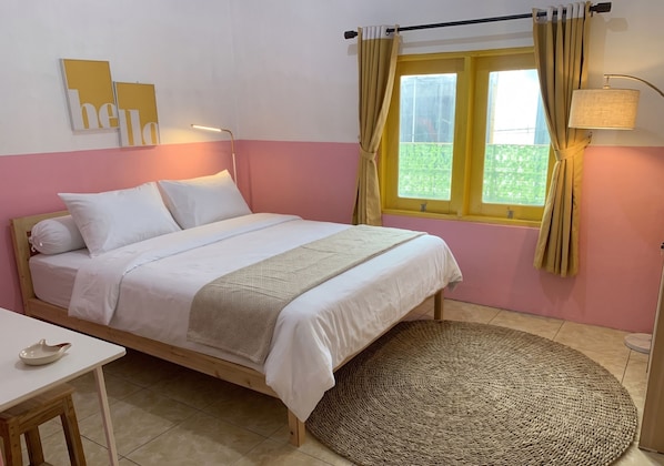 A bedroom with a bed, a bedside table, a lamp, a window with curtains, and a view of greenery outside. - Decasa Female House (Malang)