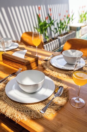 A dining table set with white plates, bowls, and cutlery, accompanied by glasses of a yellow beverage, likely wine, on a woven placemat. - Amartya F19 (Ubud)
