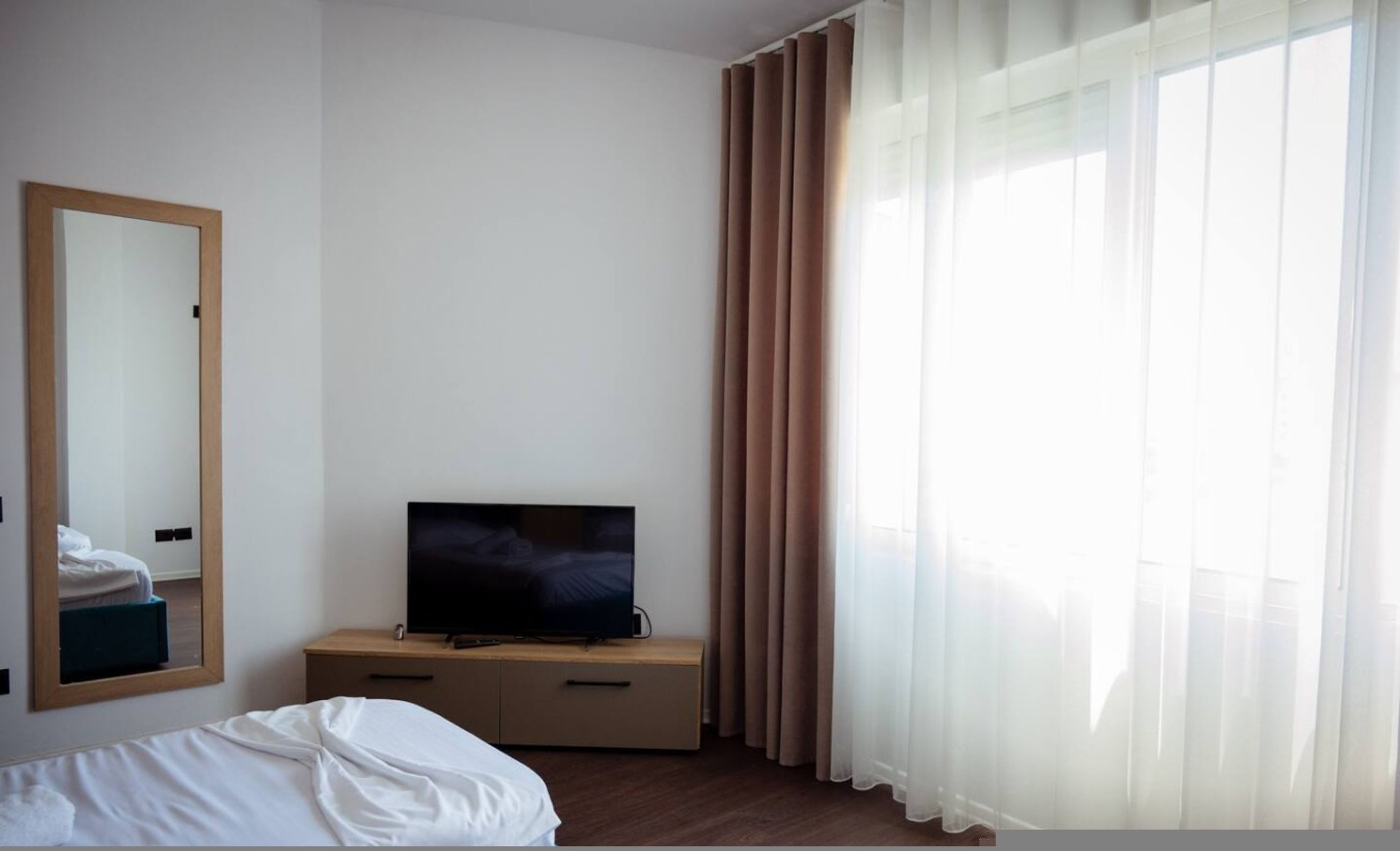 A hotel room with a bed, a television on a wooden cabinet, a mirror on the wall, and sheer curtains on the window.