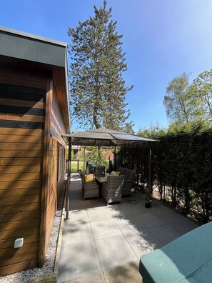 A patio area with a table and chairs, a covered seating area, and a view of a tall tree. - Cozy little chalet for a family of up to 4 people and their little rascals (Lanaken)