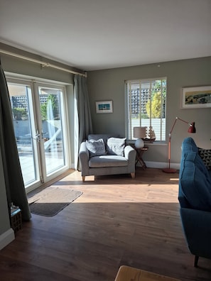 Ein Wohnzimmer mit grauem Sofa, blauem Sessel, Holzboden, einem Fenster mit Jalousien und einem gerahmten Bild an der Wand.