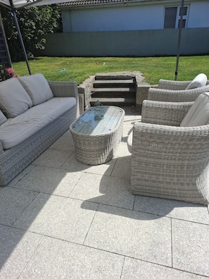 Eine Terrasseneinrichtung im Freien mit Korbmöbeln, darunter eine Couch und Stühle, sowie ein Couchtisch mit Glastischplatte.