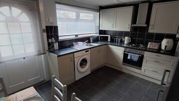 Een kleine keuken met een wasmachine, magnetron en fornuis. Er is een eettafel met borden en een stoel.