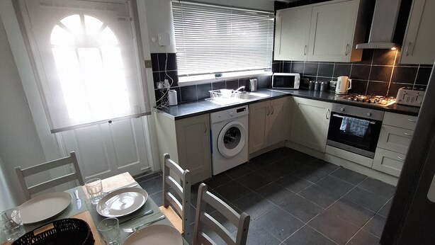 A compact kitchen with a washing machine, a dining table set with plates and cutlery, and a window with blinds. - Alice Place (Sunderland)