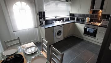 A compact kitchen with a washing machine, a dining table set with plates and cutlery, and a window with blinds.