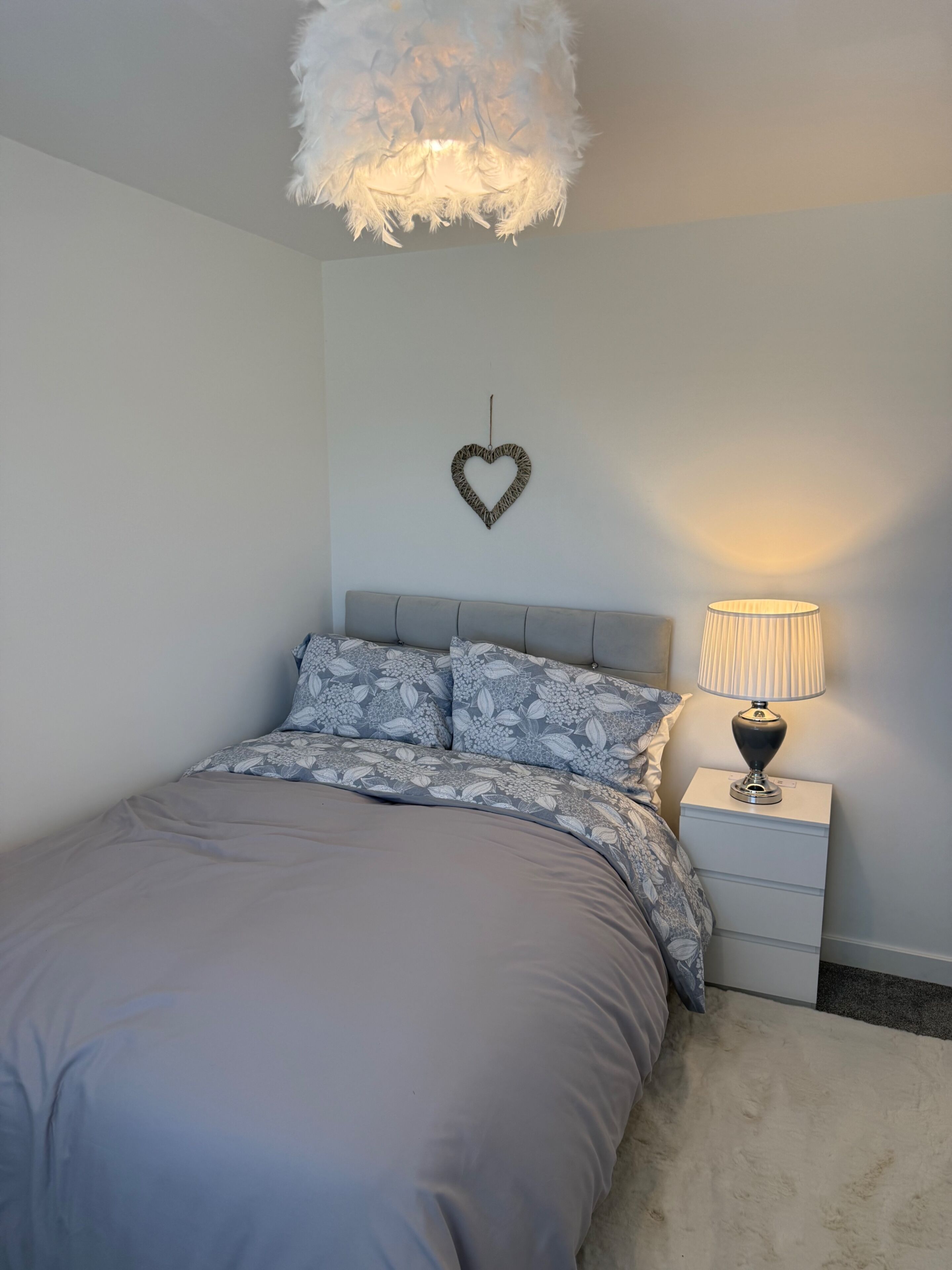 Quarto com cama, mesa de cabeceira, abajur, decoração em formato de coração e luminária de teto aconchegante.