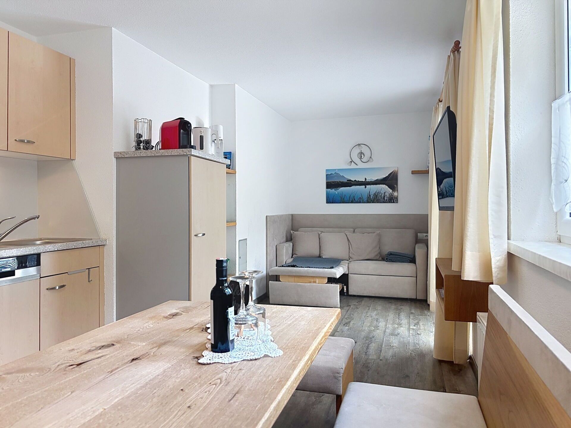 Een ruimtelijke zithoek met kitchenette, eettafel, bank en uitzicht op een meer door een raam.