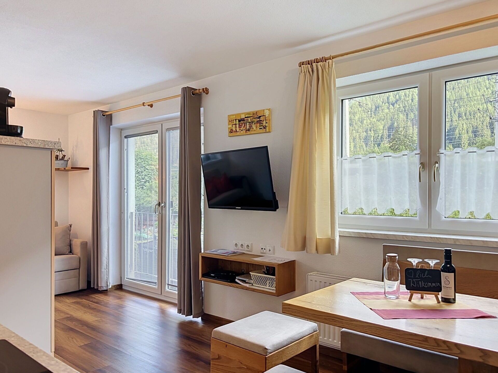 A room with a wooden floor, a TV mounted on the wall, a table with a bench, and a window with curtains.