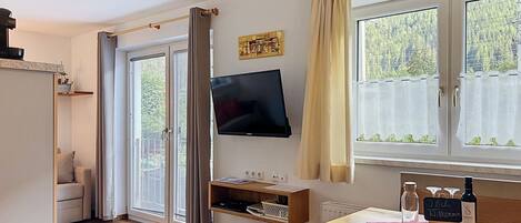 A room with a wooden floor, a TV mounted on the wall, a table with a bench, and a window with curtains.