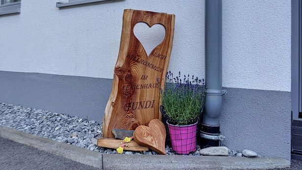 Ein hölzernes Schild mit Herz-Ausschnitt und eingraviertem Text, platziert auf einer Steinfläche mit einem Blumentopf und einem Holzgegenstand in der Nähe.