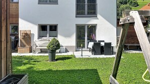 Ein zweistöckiges Gebäude mit zahlreichen Fenstern, einer Terrasse mit Gartenmöbeln und einem gepflegten Rasen.