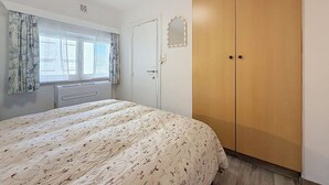 Ein Schlafzimmer mit Bett, Kleiderschrank, Fenster mit Vorhängen und Heizkörper.