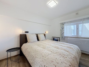 Quarto com cama, uma mesinha, janela com cortinas e radiador.