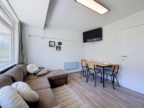 Ein modernes Wohnzimmer mit einer Couch, einem Esstisch mit Stühlen, einem an der Wand montierten Flachbildfernseher und einem Fenster mit Vorhängen.