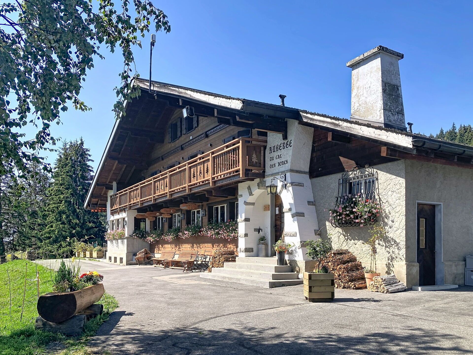 Een klassiek chalet met een houten balkon, een stenen schoorsteen en een bord met de tekst 'Anbärg des Jous'.