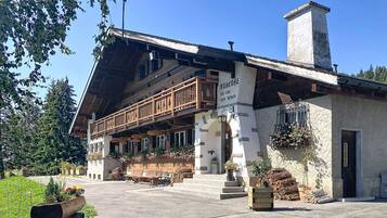Een klassiek chalet met een houten balkon, een stenen schoorsteen en een bord met de tekst 'Anbärg des Jous'.