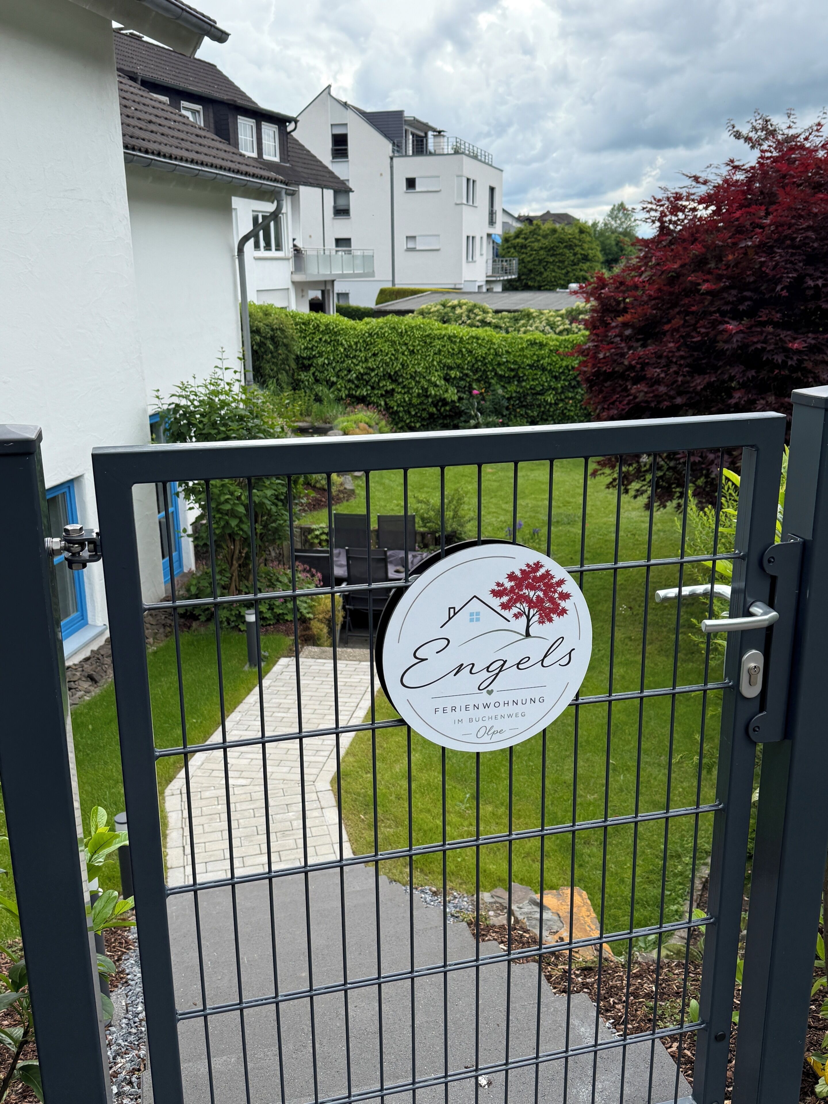 Ein verschlossenes Eingangstor mit einem Schild, auf dem 'Engels Ferienwohnung Im Buchenweg Olpe' steht.