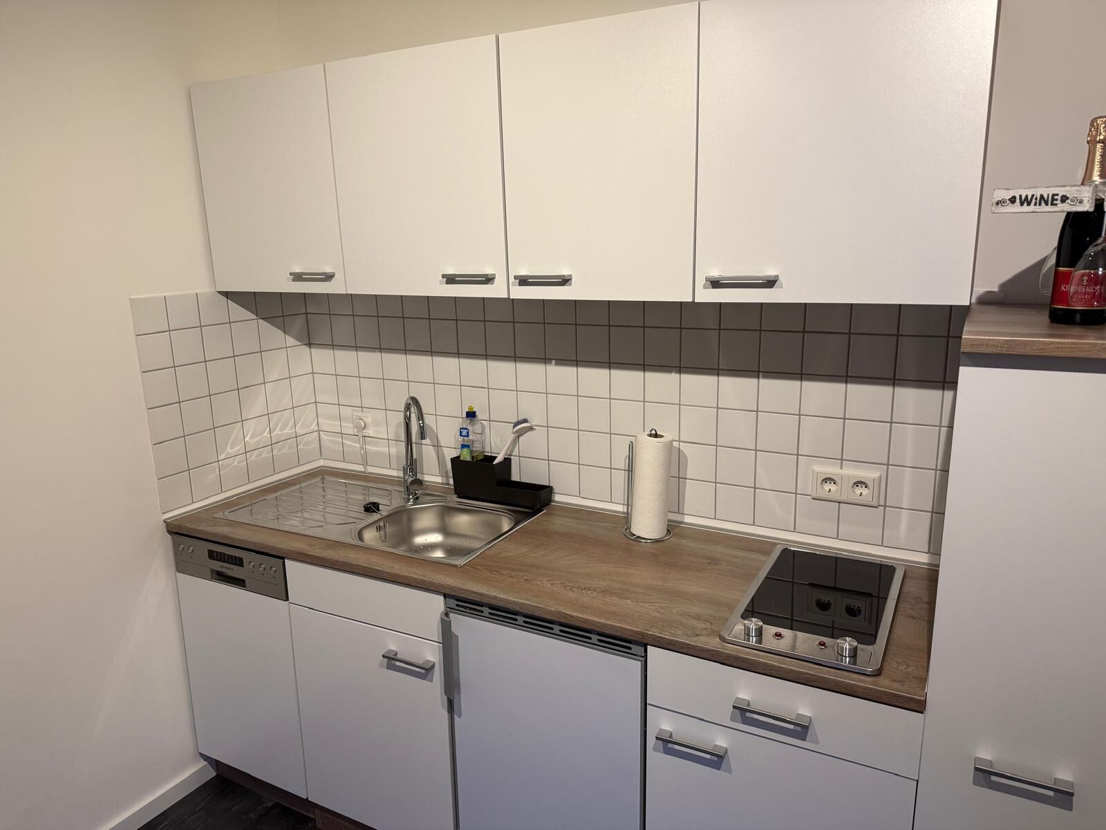 Eine moderne Küche mit weißen Schränken, einer Holz Arbeitsplatte, einem eingebauten Geschirrspüler und einer Weinflasche im Regal.