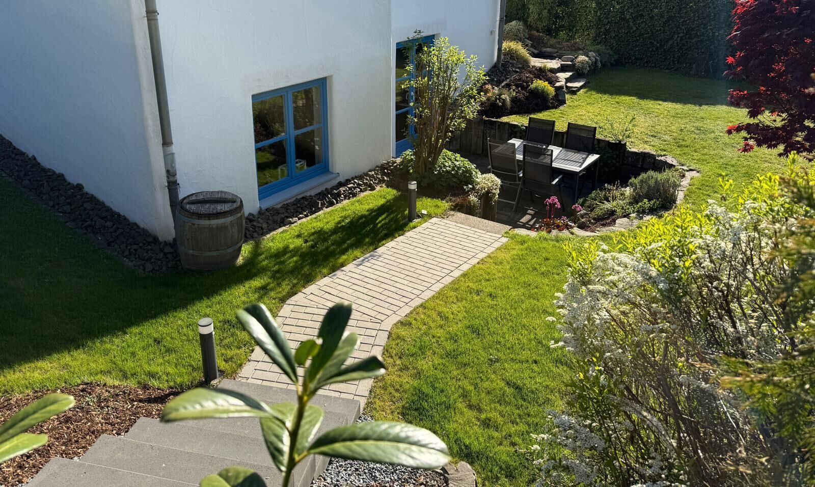 Ein Wohnhaus mit befestigter Auffahrt, einem kleinen Garten mit Tisch und Stühlen sowie verschiedenen Pflanzen und Sträuchern.