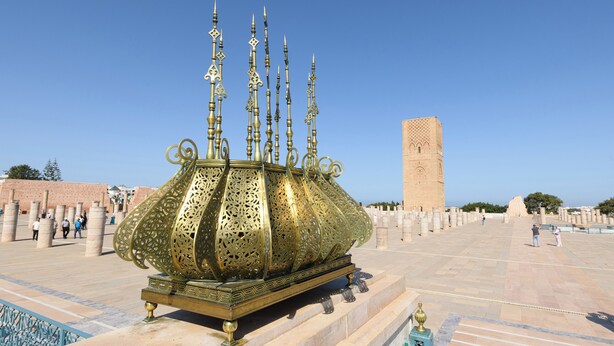 A large ornate brass candelabra with multiple candle holders, set on a platform in a courtyard with a tall tower in the background.