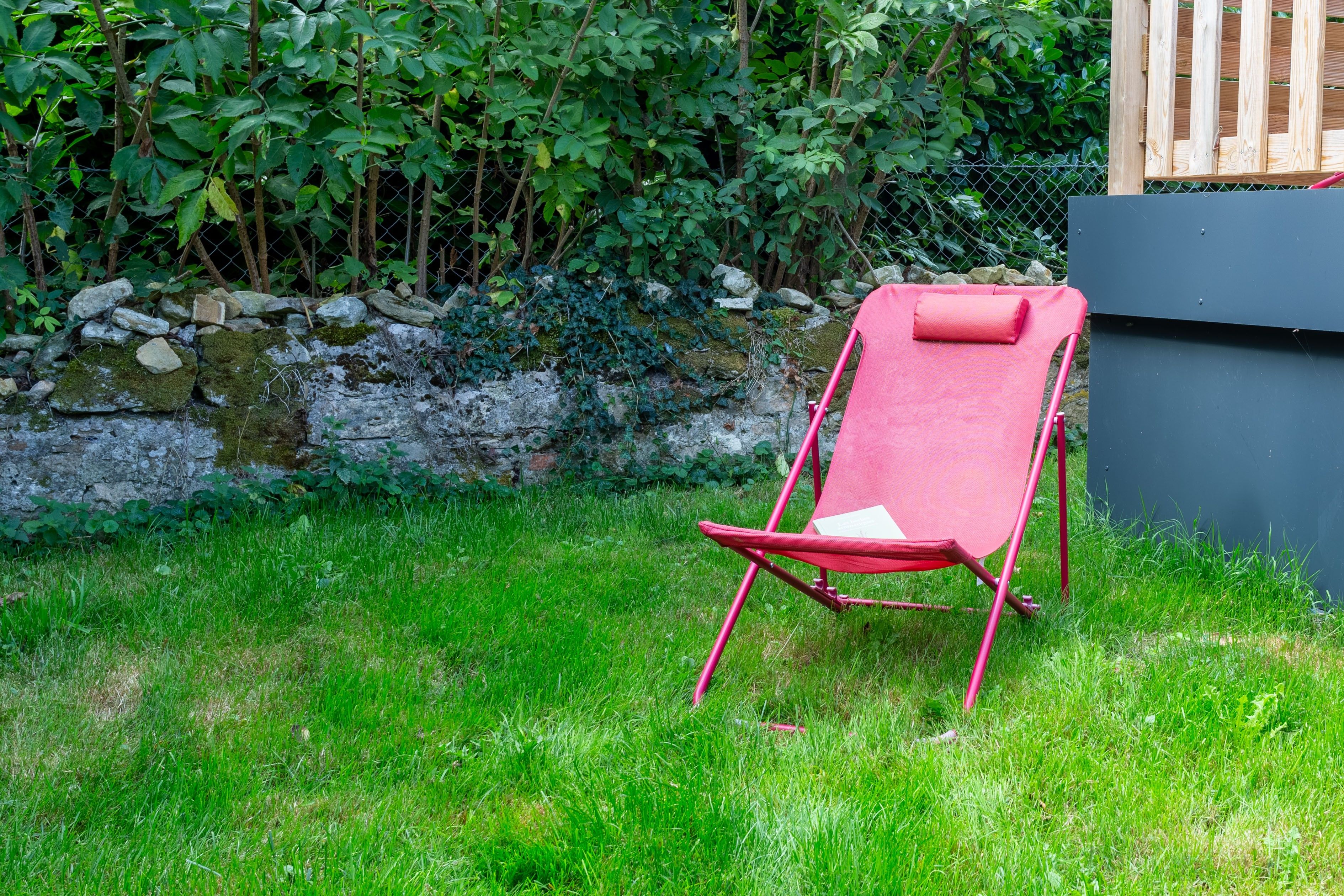 A red lounge chair in a grassy area with a stone wall and dense foliage in the background.
