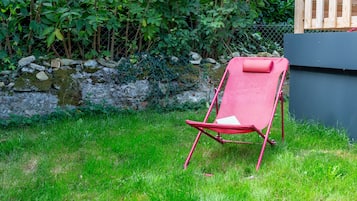 A red lounge chair in a grassy area with a stone wall and dense foliage in the background.
