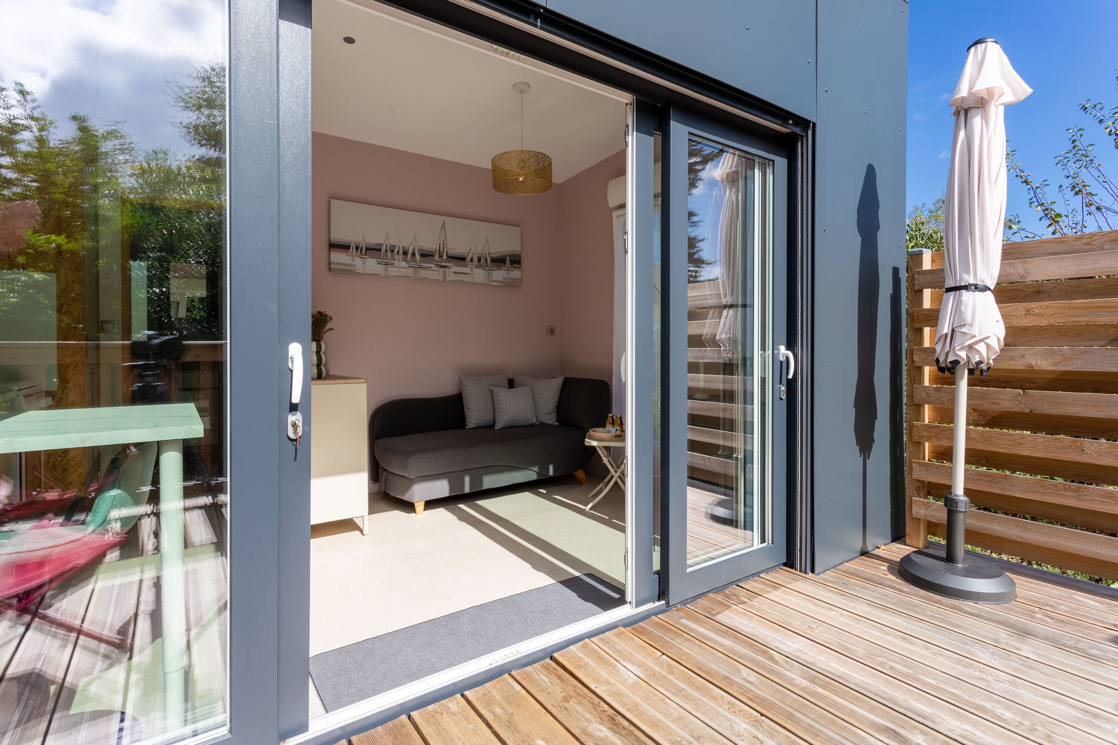 Una sala de estar moderna con un sofá, un cuadro y un ventanal que da a un área exterior con terraza de madera y una sombrilla.