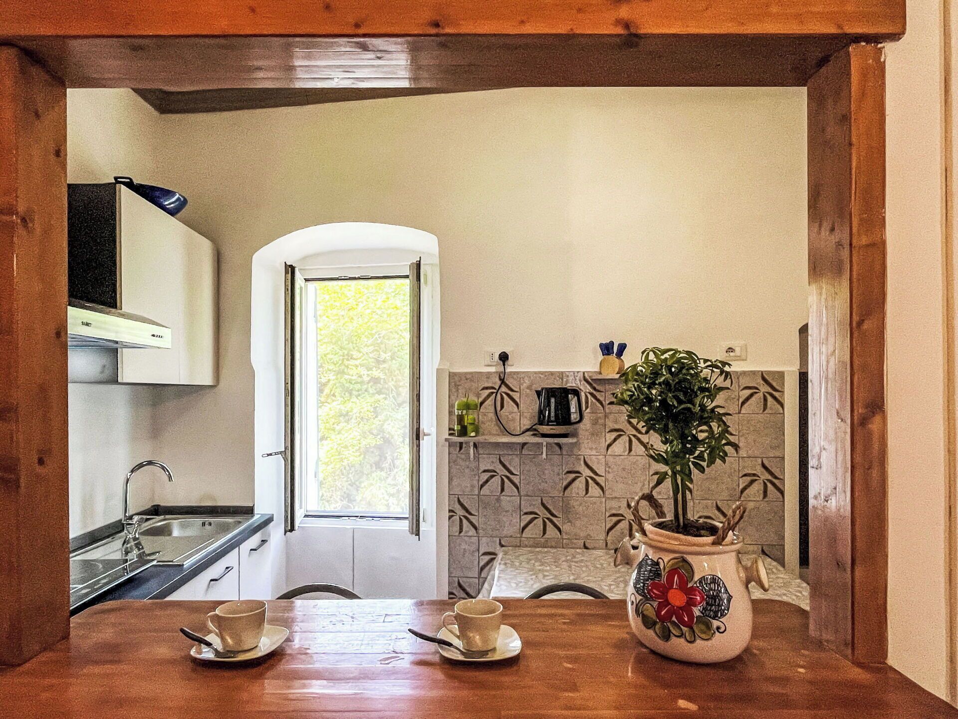 Een keuken met een houten tafel, twee kopjes, een plant in een decoratieve pot en een raam met uitzicht op bomen.