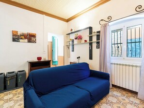 Ein Zimmer mit einem blauen Sofa, einer Holzbalkendecke, einem Heizkörper und einem Fenster mit Metallgitter.