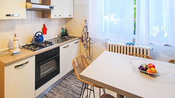 Une cuisine moderne équipée d’une cuisinière à gaz, d’un four et de meubles blancs. On y trouve une table à manger avec un panier de fruits et une chaise en osier.
