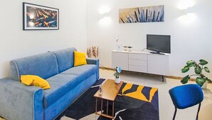 Un salon moderne avec un canapé bleu, une table basse, un téléviseur et des œuvres d’art accrochées aux murs.
