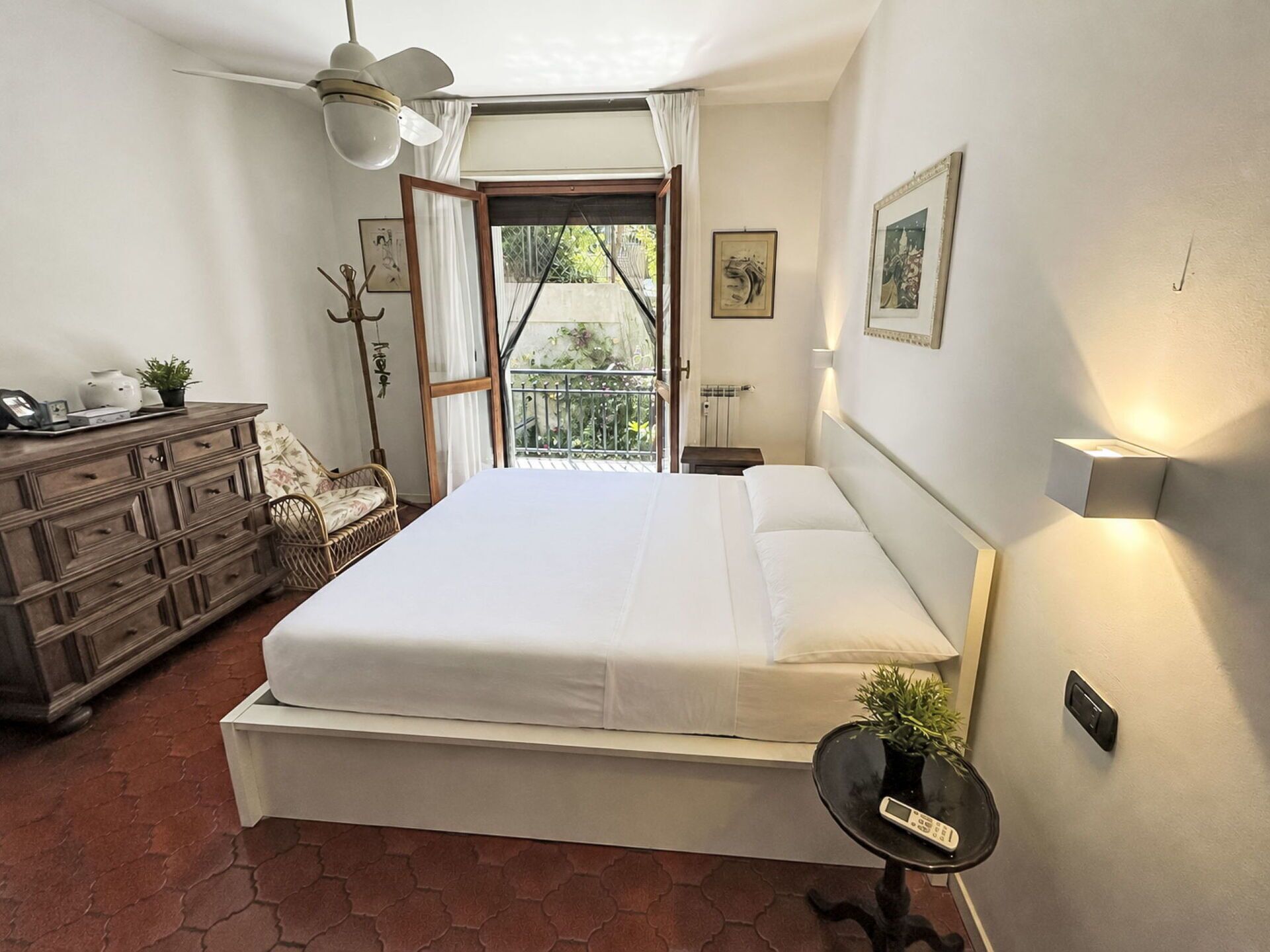 Ein Schlafzimmer mit einem großen Bett, einem Schminktisch, einem Sessel, einem kleinen Tisch mit Pflanze und einem Balkon mit Aussicht.