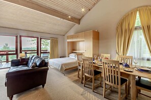 Ein Zimmer mit einem Bett, einer Schlafcouch, einem Esstisch mit Stühlen und einem großen Fenster mit Vorhängen.