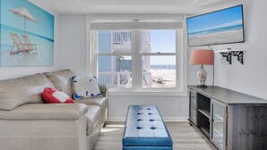 A living room with a sofa, a bench, a TV, and a window with a view of the beach. - Peaceful Pearl (North Topsail Beach)