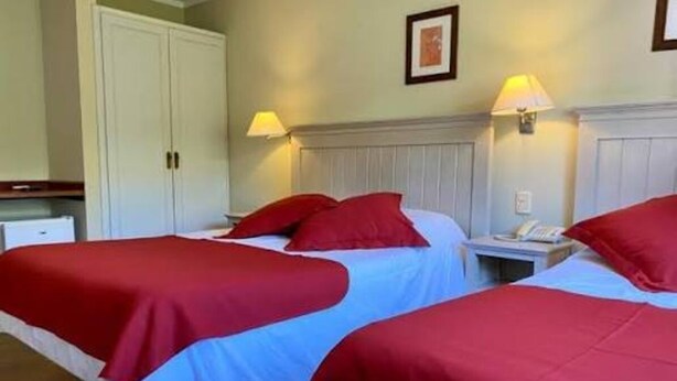 A hotel room with two beds, each with red bedding, a nightstand with a lamp, and a wardrobe.