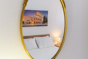 Ein Bett mit weißen Leinen, ein Nachttisch mit Lampe und ein gerahmtes Bild des Kolosseums an der Wand.